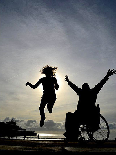 figure in wheelchair with arms in air anda second figure leaping into air
